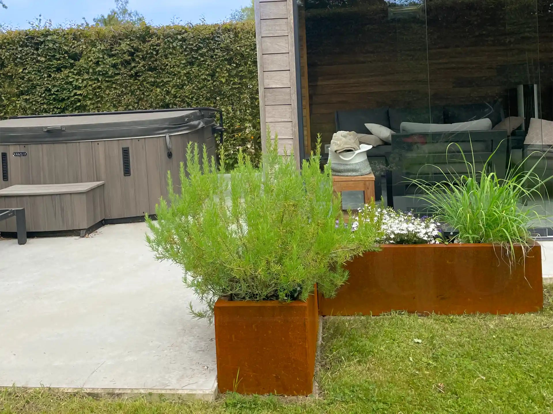 Tuin met twee cortenstalen plantenbakken van Adeqo, beplant met lavendel, siergras en witte bloemen, naast een terras met jacuzzi en zithoek.