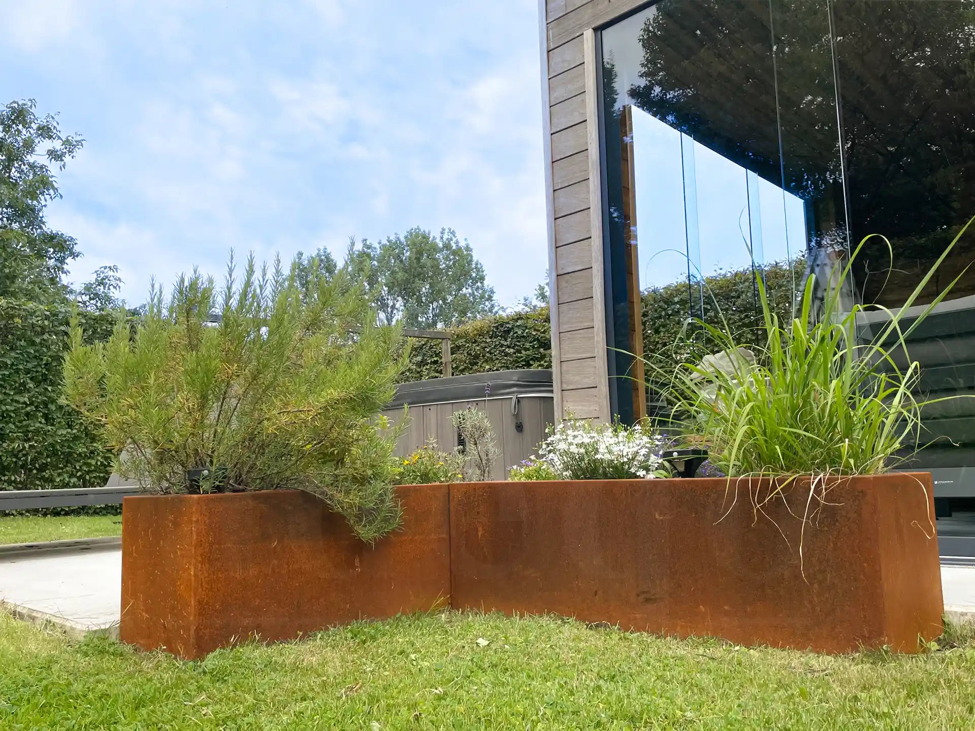 Tuinhoek met twee cortenstalen plantenbakken van Adeqo, beplant met rozemarijn, siergras en witte bloemen, naast een modern huis met houten gevel en grote ramen.