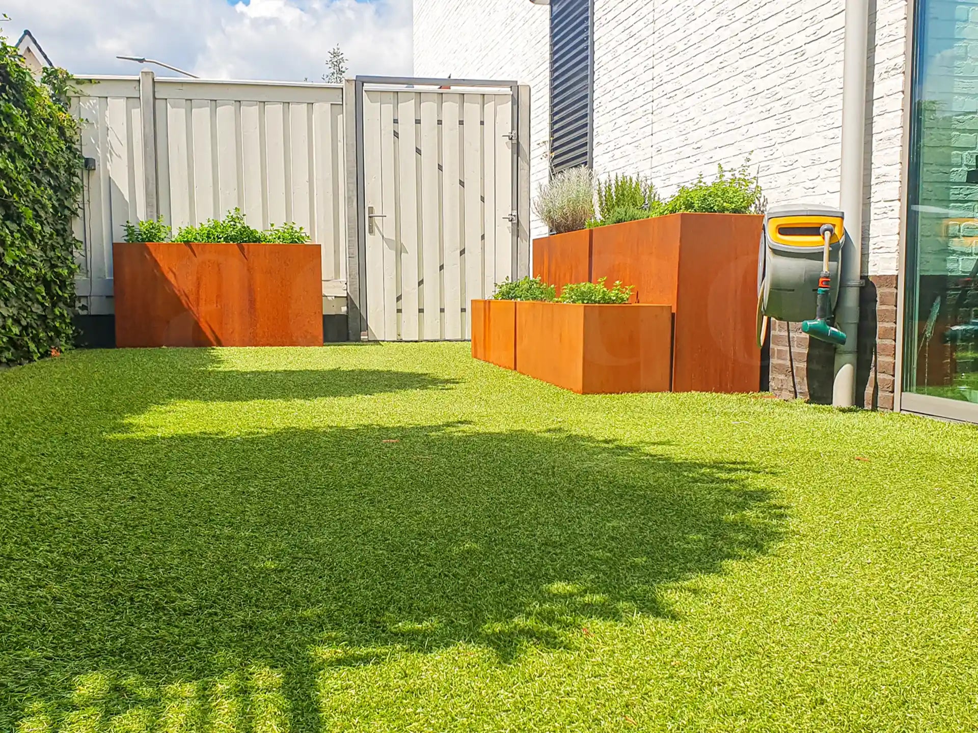 Moderne tuin met kunstgras en meerdere cortenstalen plantenbakken van Adeqo, beplant met groene planten en kruiden, geplaatst langs een witte schutting.