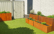 Tuin met kunstgras en cortenstalen plantenbakken van Adeqo, gevuld met kruidenplanten en lavendel, geplaatst langs een witte schutting en muur.