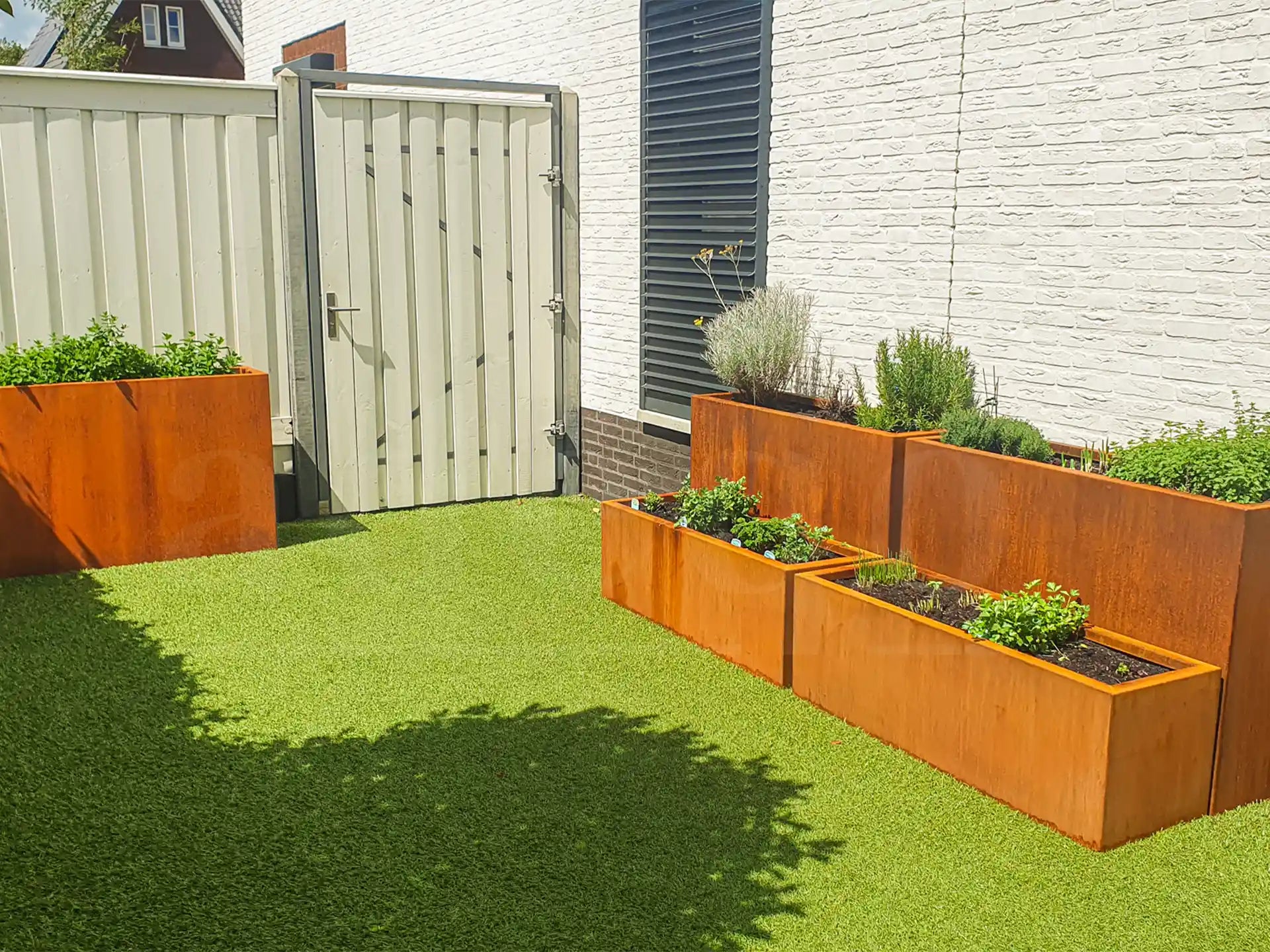 Tuin met kunstgras en cortenstalen plantenbakken van Adeqo, gevuld met kruidenplanten en lavendel, geplaatst langs een witte schutting en muur.