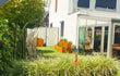 Moderne tuin met meerdere cortenstalen plantenbakken van Adeqo op kunstgras, beplant met siergrassen en bloemen, naast een loungebank, hangstoel en een serre.