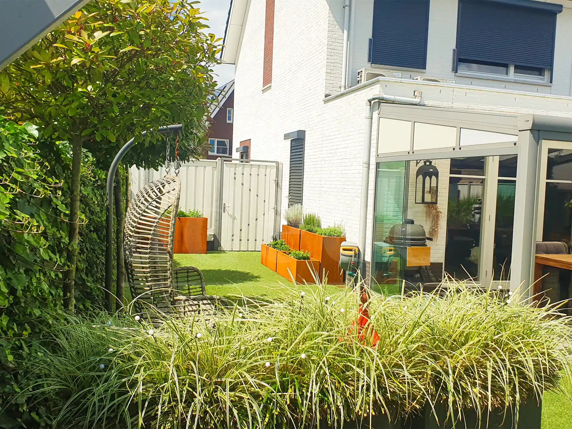 Moderne tuin met meerdere cortenstalen plantenbakken van Adeqo op kunstgras, beplant met siergrassen en bloemen, naast een loungebank, hangstoel en een serre.