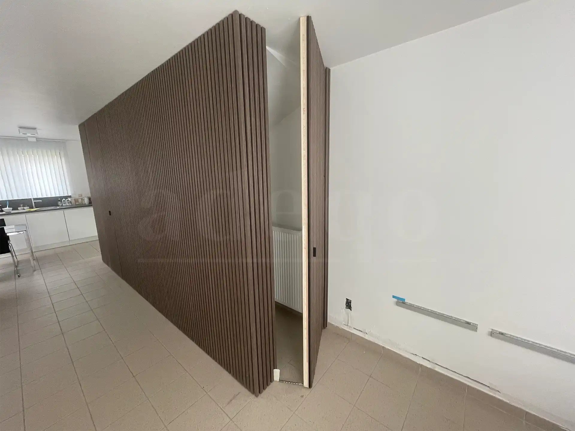 Openstaande verborgen deur in een wand bekleed met Adeqo akoestische panelen in Donker Walnoot, met zicht op radiator en keukenruimte op de achtergrond.