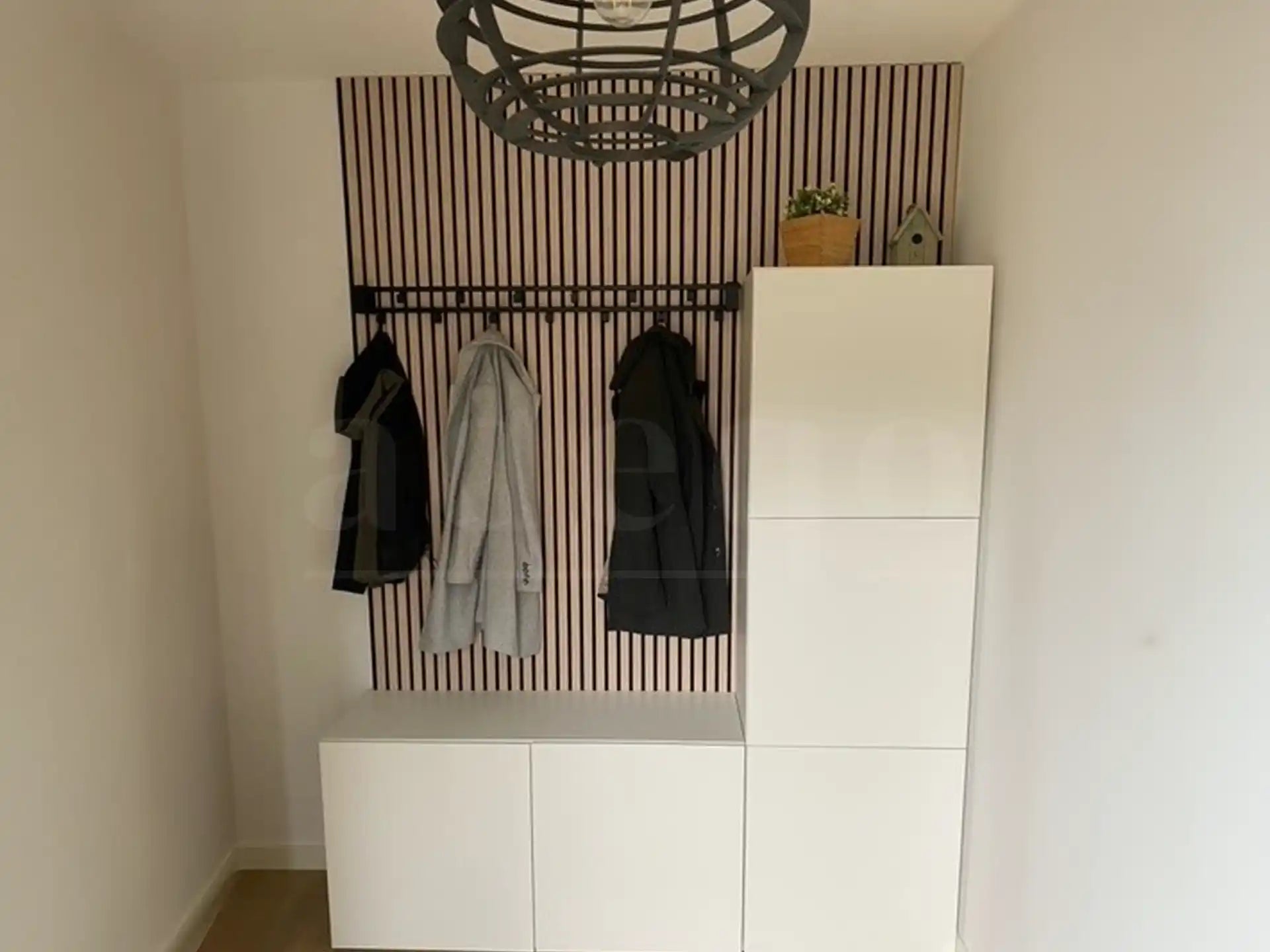 Hal met witte kasten en kapstok tegen een wand met Adeqo akoestische panelen in Wit Eiken, voorzien van jassen en een decoratief plantje.