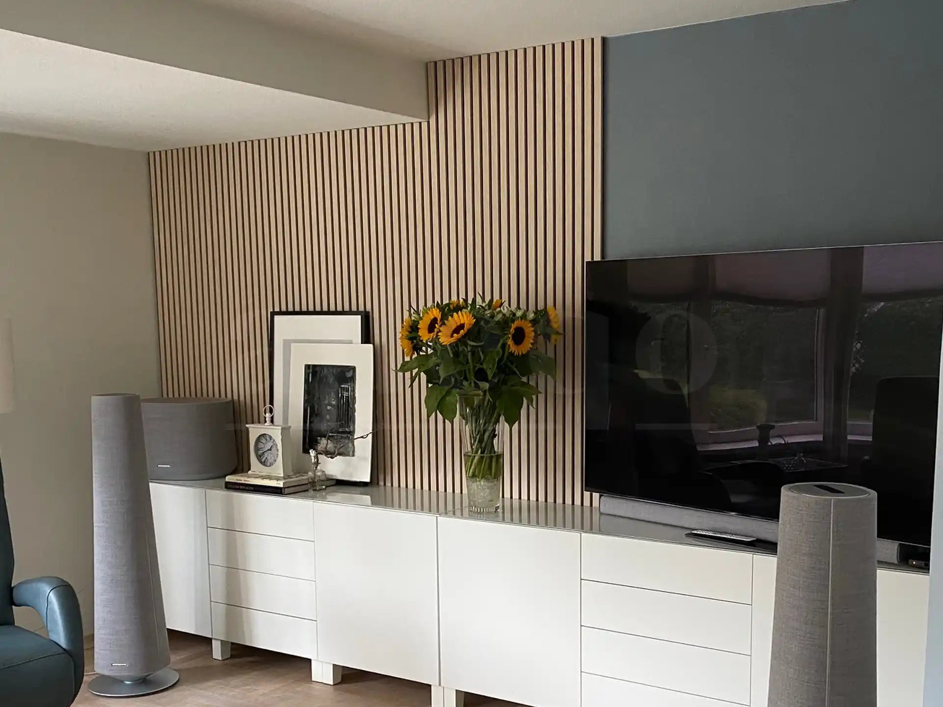 Woonkamer met Adeqo akoestische panelen in Wit Eiken achter een wit dressoir, versierd met zonnebloemen, schilderijen en een televisie.