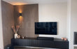 Woonkamerhoek met Adeqo akoestisch wandpaneel in Licht Walnoot, zwart tv-meubel en moderne wandlamp.