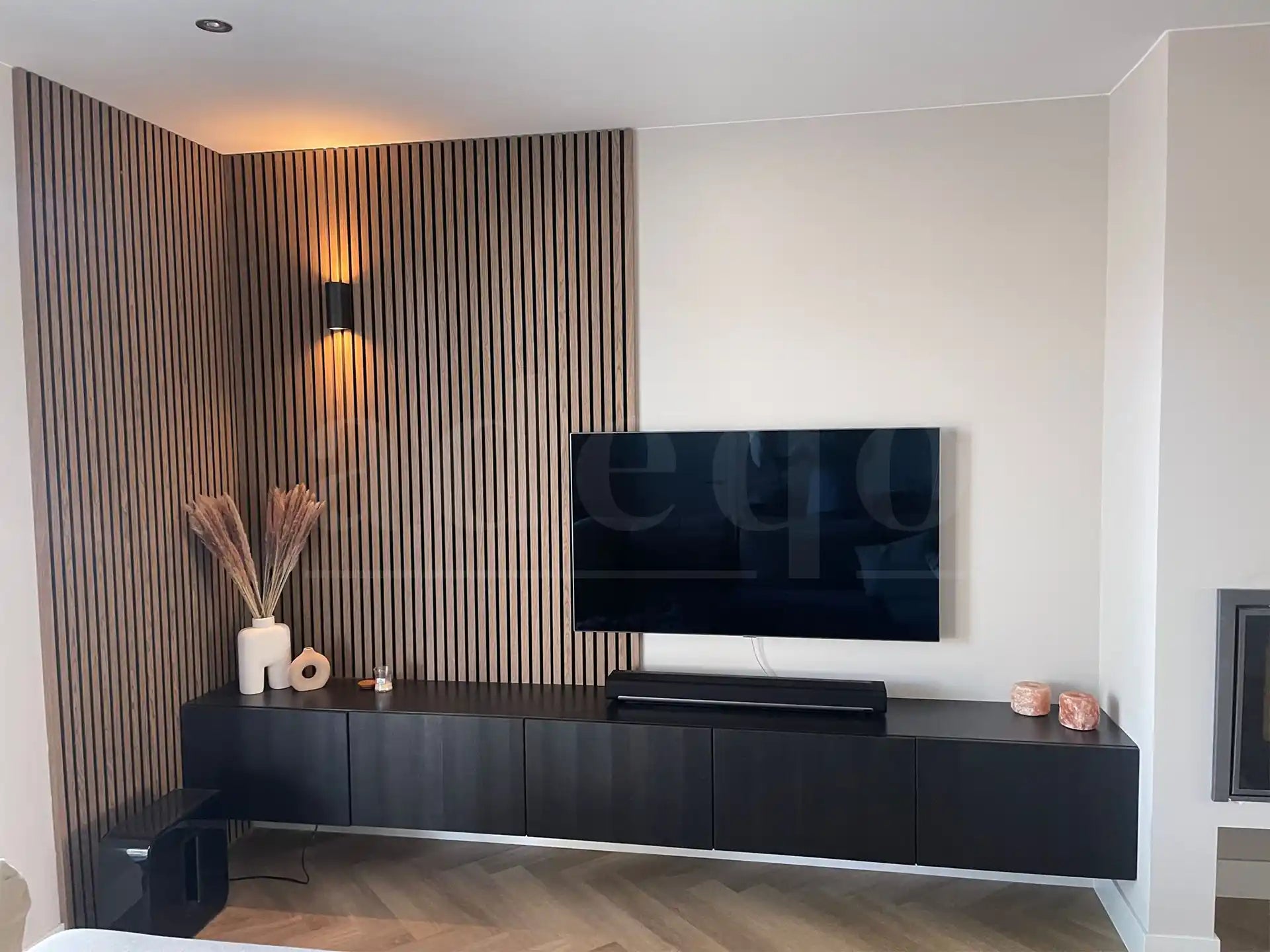 Woonkamerhoek met Adeqo akoestisch wandpaneel in Licht Walnoot, zwart tv-meubel en moderne wandlamp.