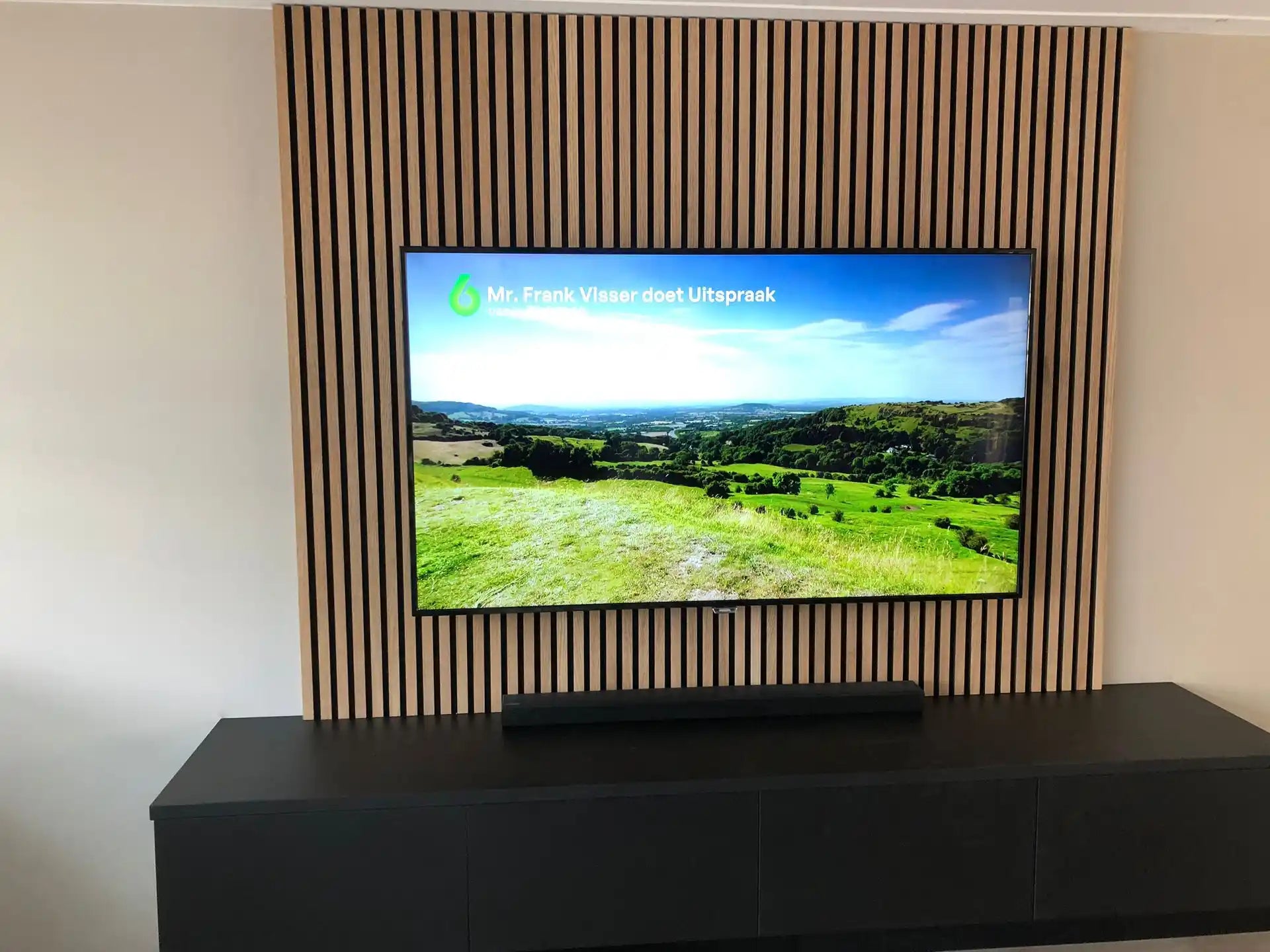 Tv-wand met Adeqo akoestisch wandpaneel in Natuurlijk Eiken achter een zwart tv-meubel.