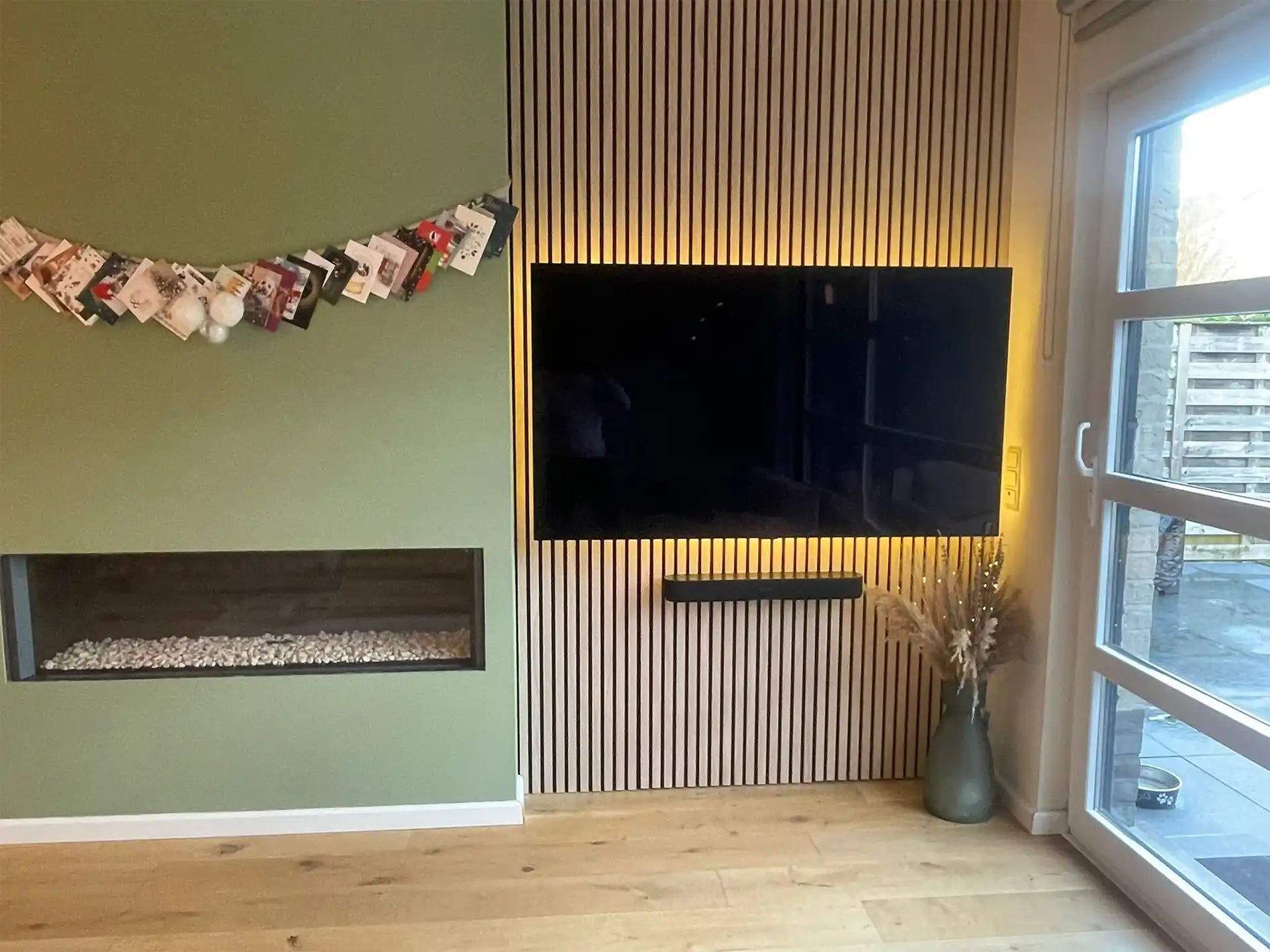 Tv-wand met Adeqo akoestisch wandpaneel in Natuurlijk Eiken en geïntegreerde verlichting, naast groene wand met haard.