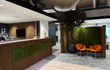 Moderne kantoorbalie met Bruine Eiken akoestische wandpanelen van Adeqo en een groen mosaccent, naast een zithoek met oranje stoelen en een grote decoratieve boom.