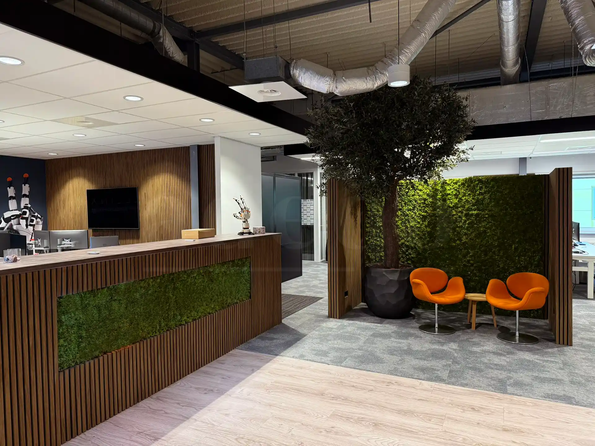 Moderne kantoorbalie met Bruine Eiken akoestische wandpanelen van Adeqo en een groen mosaccent, naast een zithoek met oranje stoelen en een grote decoratieve boom.