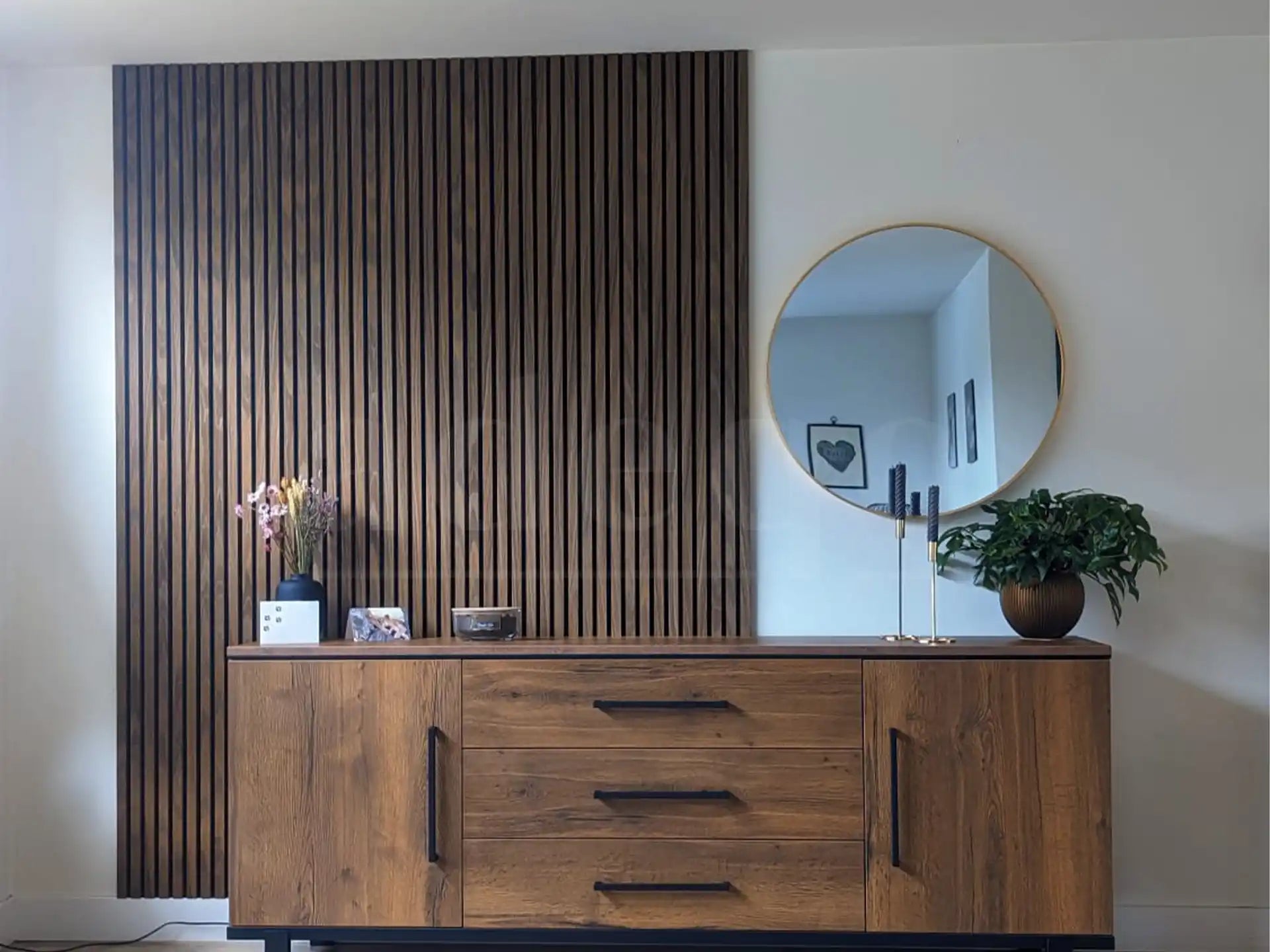 Moderne woonkamer met Adeqo akoestische wandpanelen in Bruin Eiken achter een houten dressoir, gedecoreerd met planten, kaarsen en een ronde spiegel.