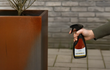 Hand die Adeqo cortenstaal roestversneller sprayt op een grote cortenstalen plantenbak met jonge boom, tegen een achtergrond van grijze stenen muur en bestrating.
