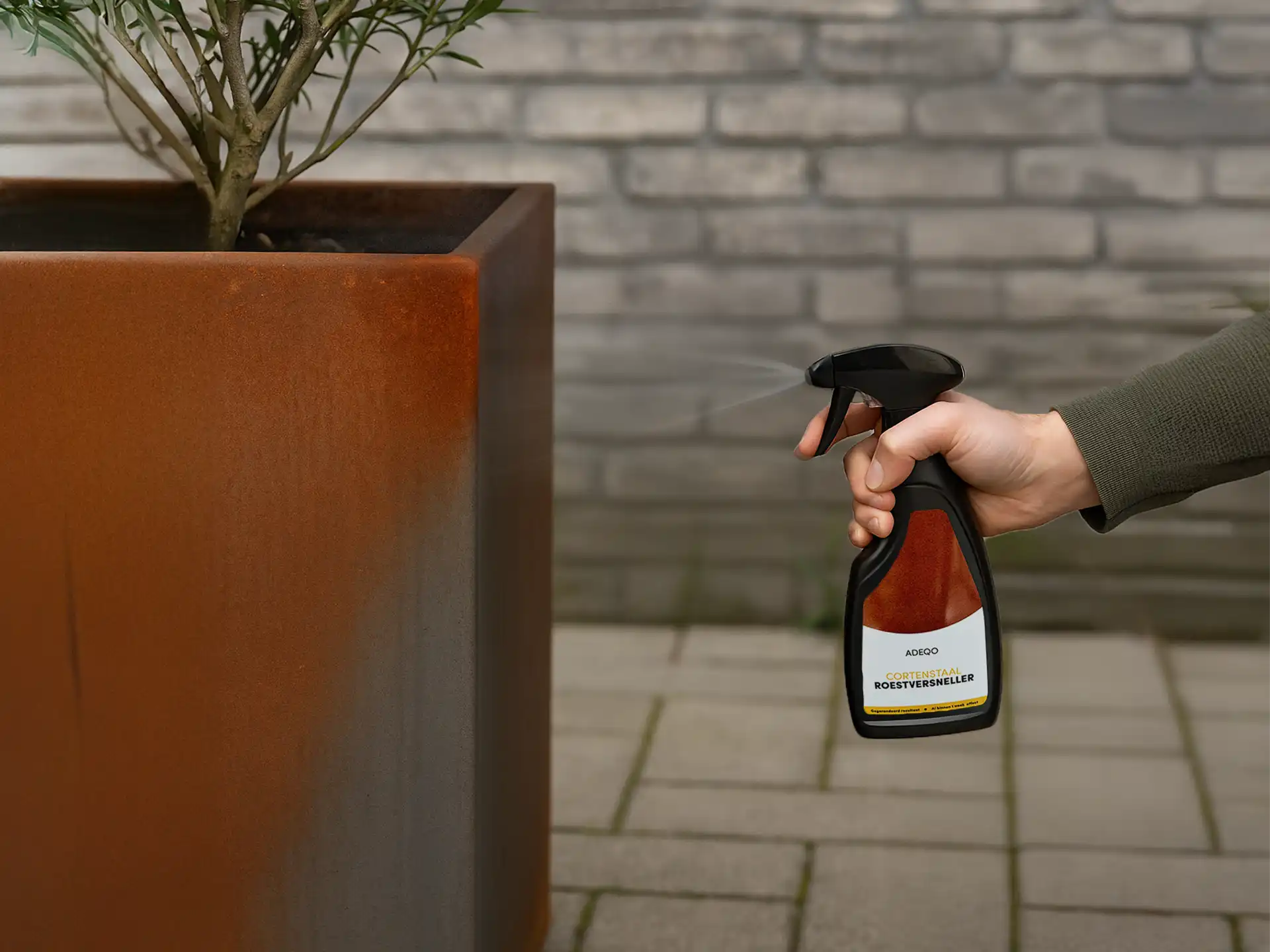 Hand die Adeqo cortenstaal roestversneller sprayt op een grote cortenstalen plantenbak met jonge boom, tegen een achtergrond van grijze stenen muur en bestrating.