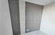 Strakke wandbekleding met Adeqo akoestische wandpanelen in Grijs Eiken, gecombineerd met een middenstrook van licht natuursteen, geplaatst in een moderne ruimte met glanzende witte vloer.