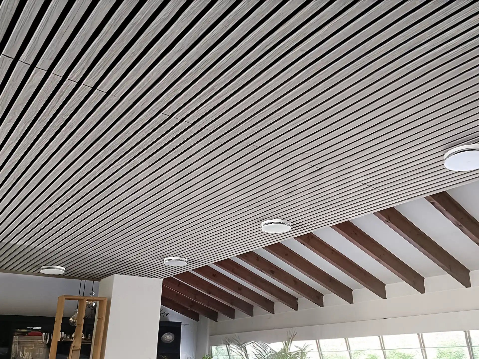 Modern plafond afgewerkt met Adeqo akoestische wandpanelen in Grijs Eiken, voorzien van geïntegreerde ronde inbouwspots en zichtbare houten dakbalken.