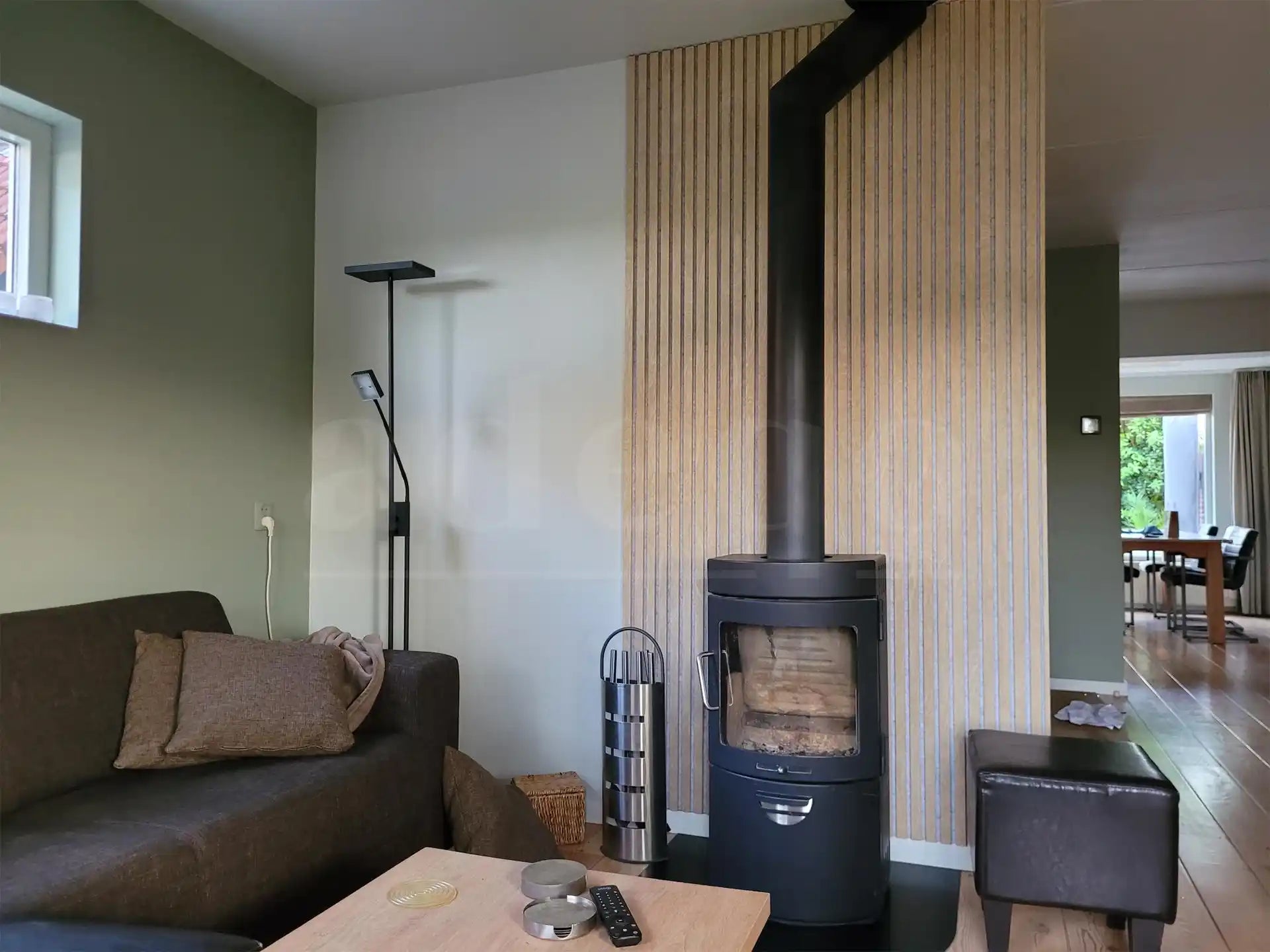 Woonkamer met zwarte houtkachel tegen een wand bekleed met Adeqo akoestische wandpanelen in Natuurlijk Eiken met grijs vilt, voor een warme en stijlvolle uitstraling.