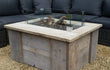 Adeqo Steigerhout Vuurtafel Firenze in vierkante uitvoering, gemaakt van oud steigerhout met ingebouwde brander en glasombouw, geplaatst op een terras met loungeset.