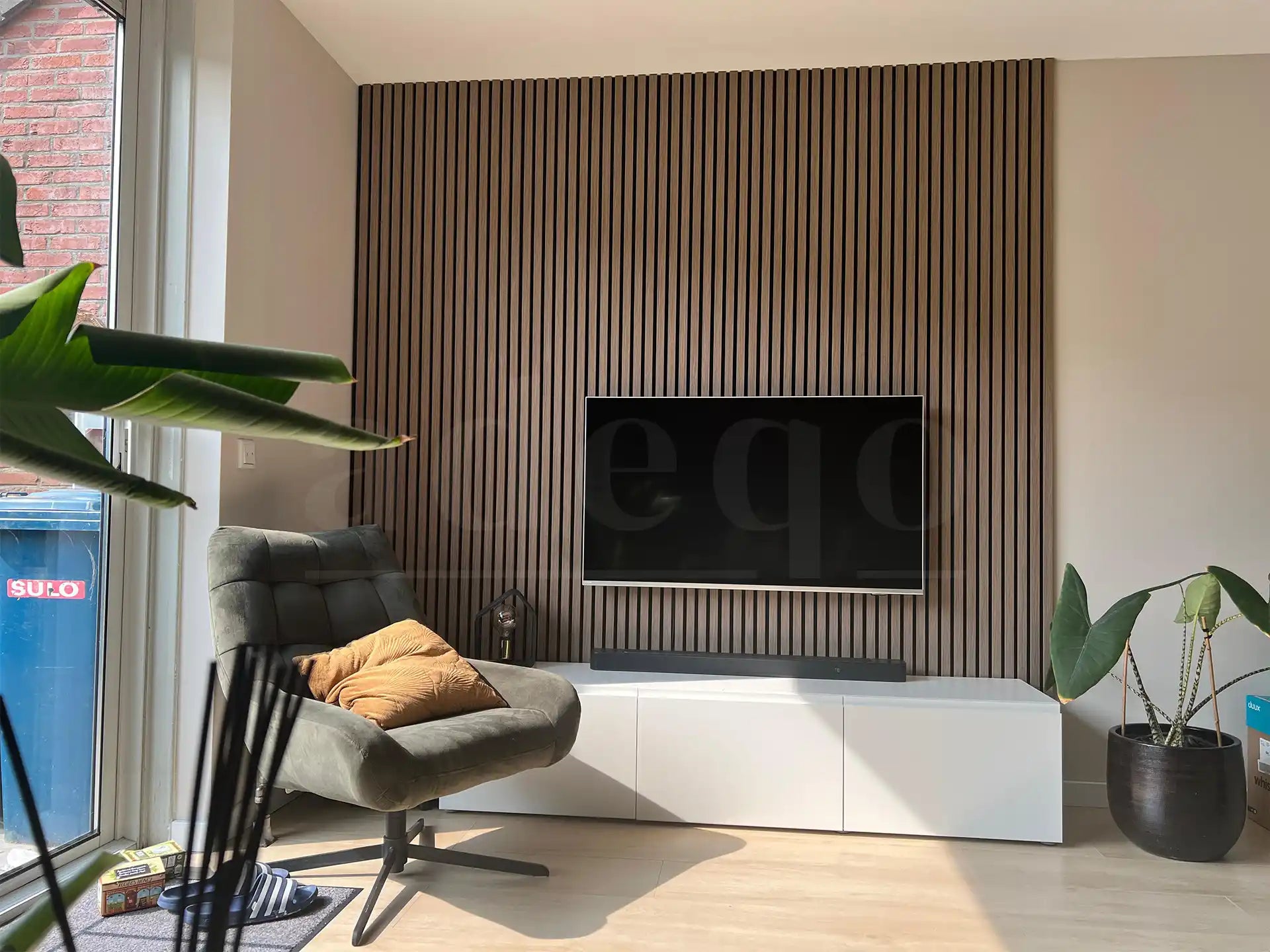 Woonkamer met moderne tv-wand bekleed met Adeqo akoestische wandpanelen in Verouderd Eiken, gecombineerd met een strak wit tv-meubel en een comfortabele fauteuil.