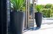 Twee ronde Adezz Canna polyester plantenbakken in donkergrijs met groene planten op een terras.