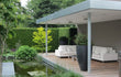 Grote ronde Adezz Canna polyester plantenbak in donkergrijs naast een moderne overkapping met vijver.