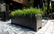 Grote zwarte Adezz Carrez aluminium plantenbak met poten naast een horecaterras, gevuld met groene beplanting.