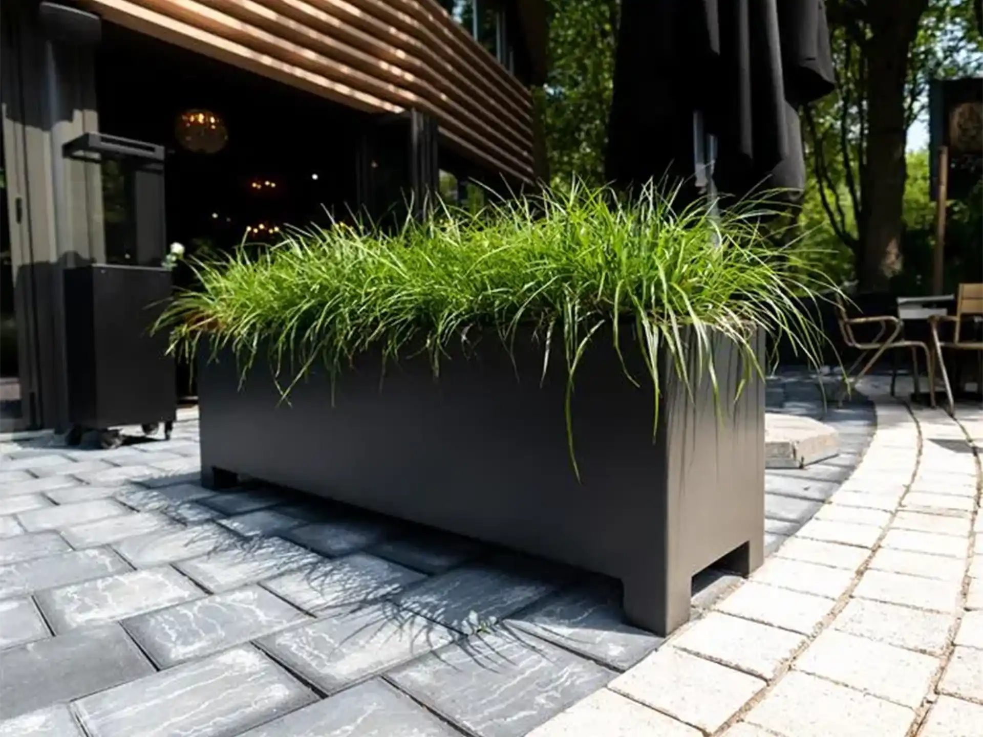 Grote zwarte Adezz Carrez aluminium plantenbak met poten naast een horecaterras, gevuld met groene beplanting.