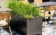 Zwarte Adezz Carrez aluminium plantenbak met poten, gevuld met siergrassen, geplaatst op een terras met stoelen.