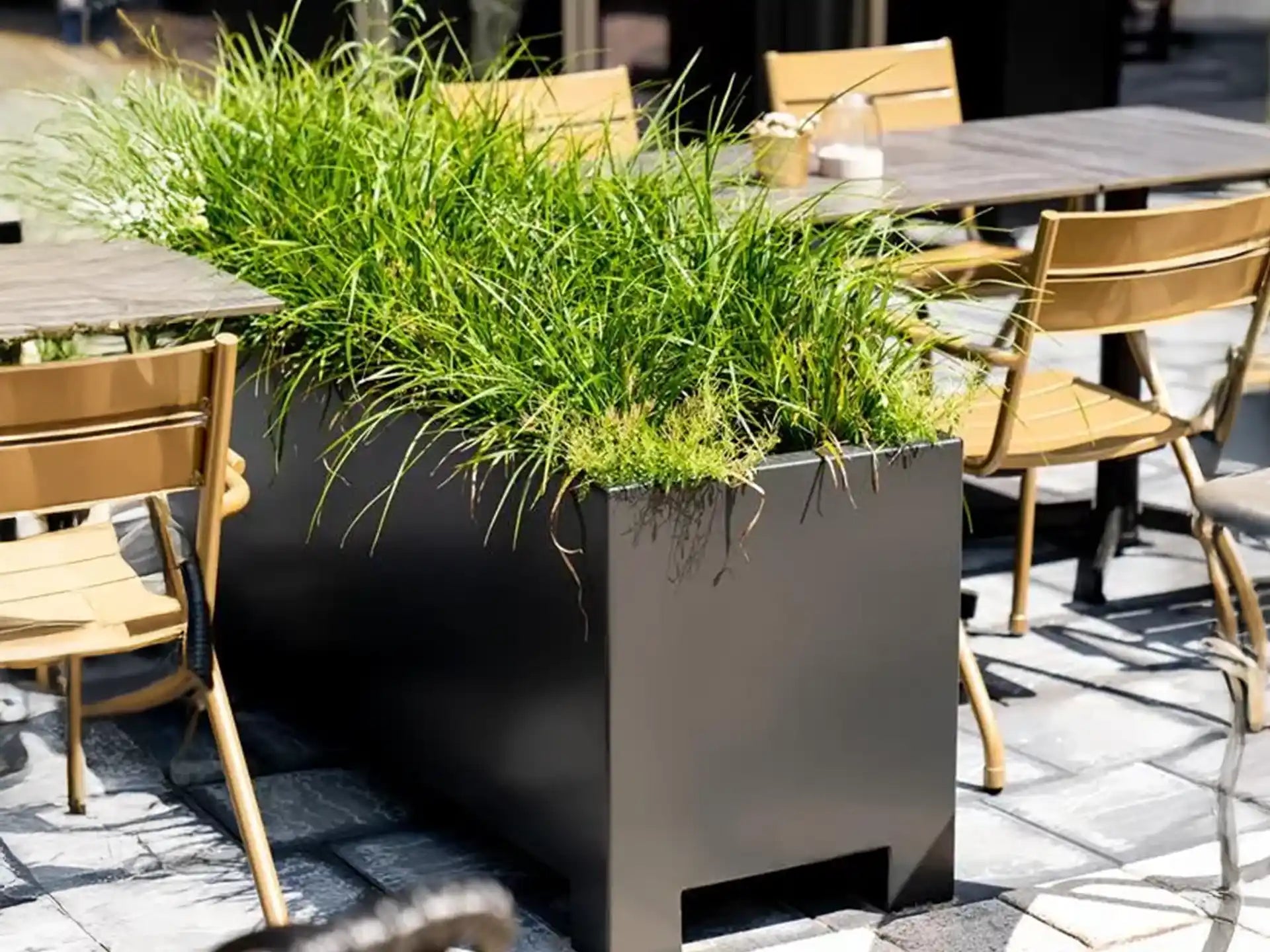 Zwarte Adezz Carrez aluminium plantenbak met poten, gevuld met siergrassen, geplaatst op een terras met stoelen.