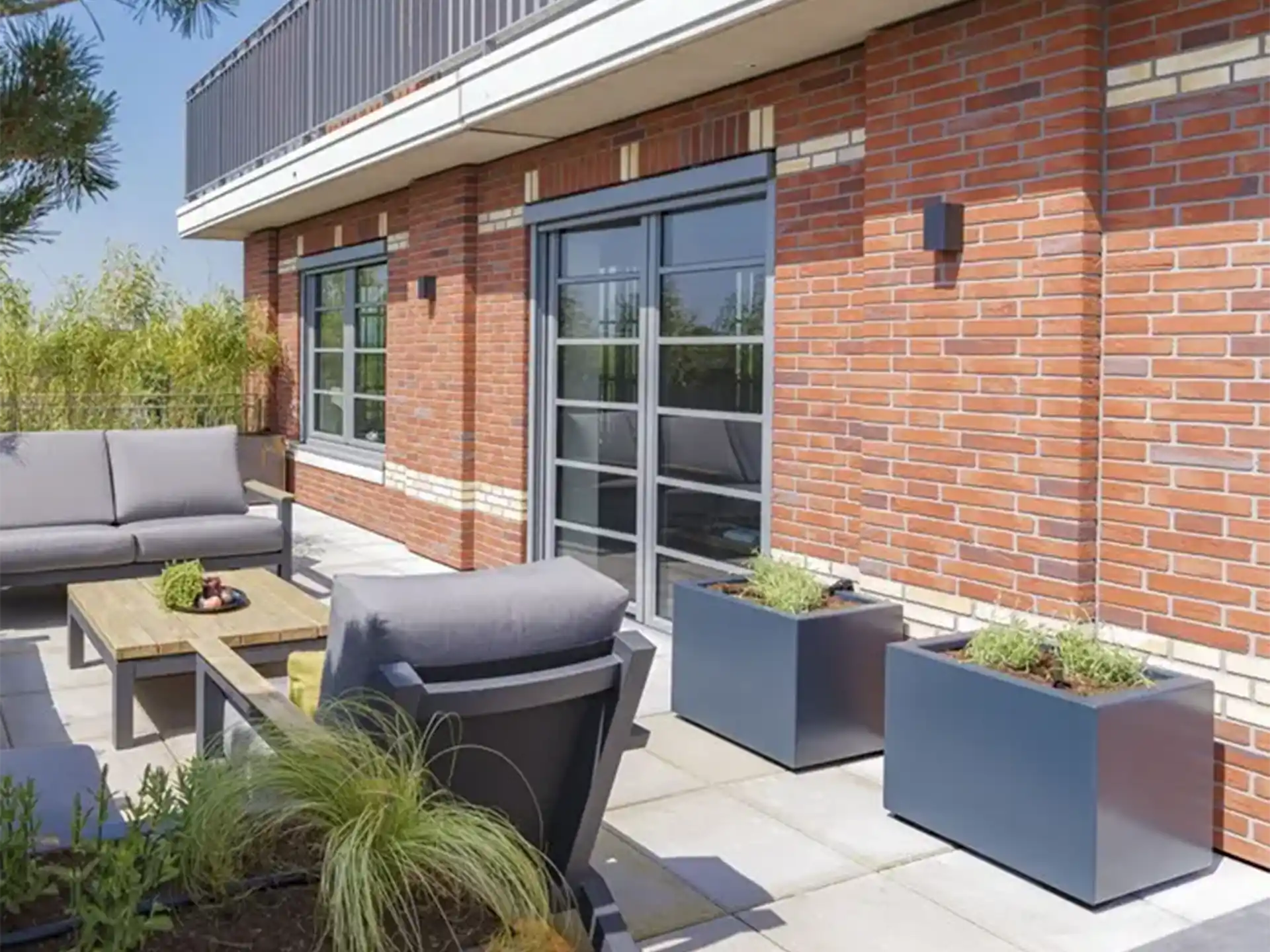 Meerdere rechthoekige Adezz Carrez aluminium plantenbakken met wielen naast een moderne woning en lounge set.