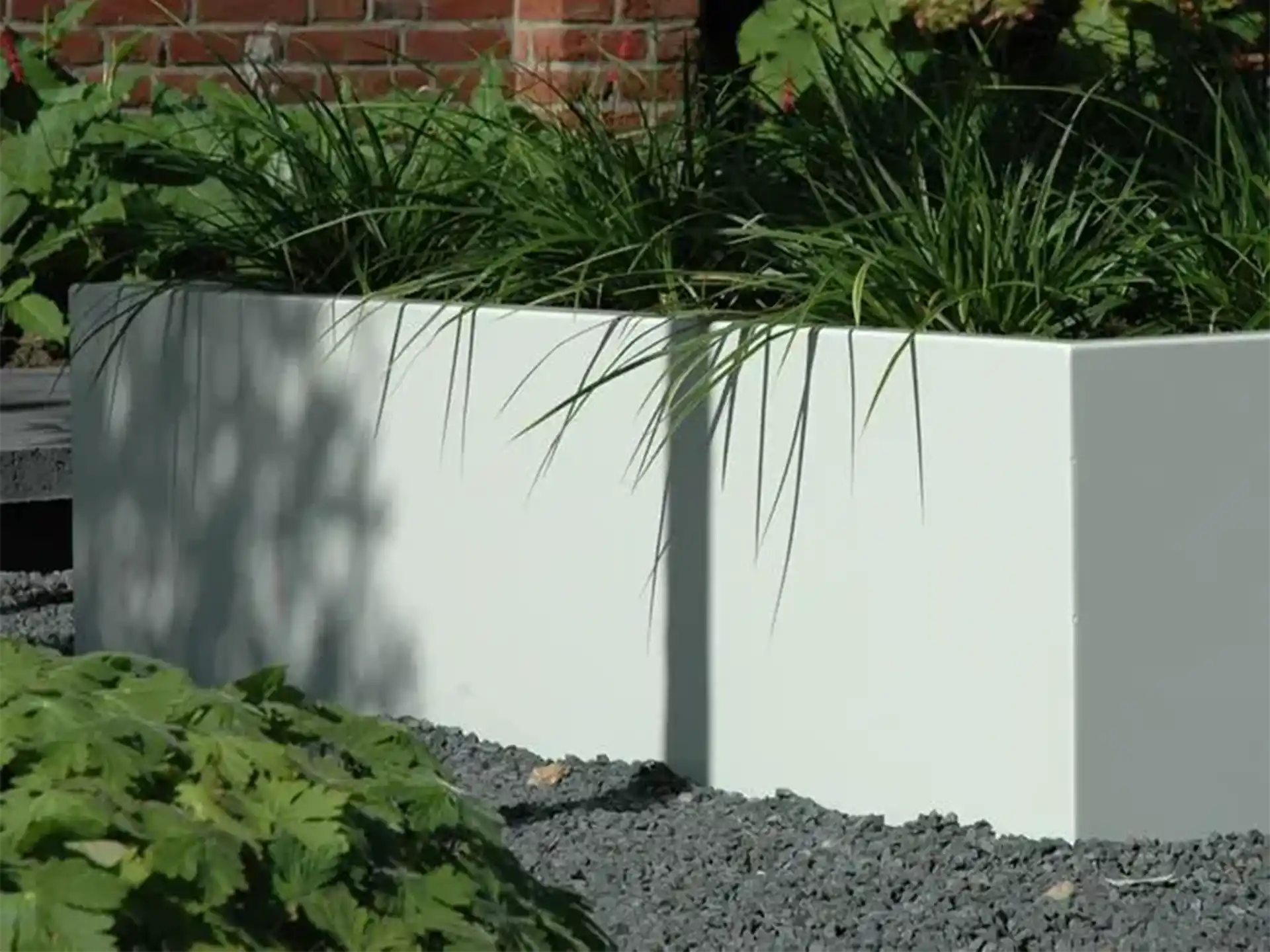 Witte Adezz Carrez aluminium plantenbak zonder bodem in een tuin, beplant met siergrassen en omgeven door grind.