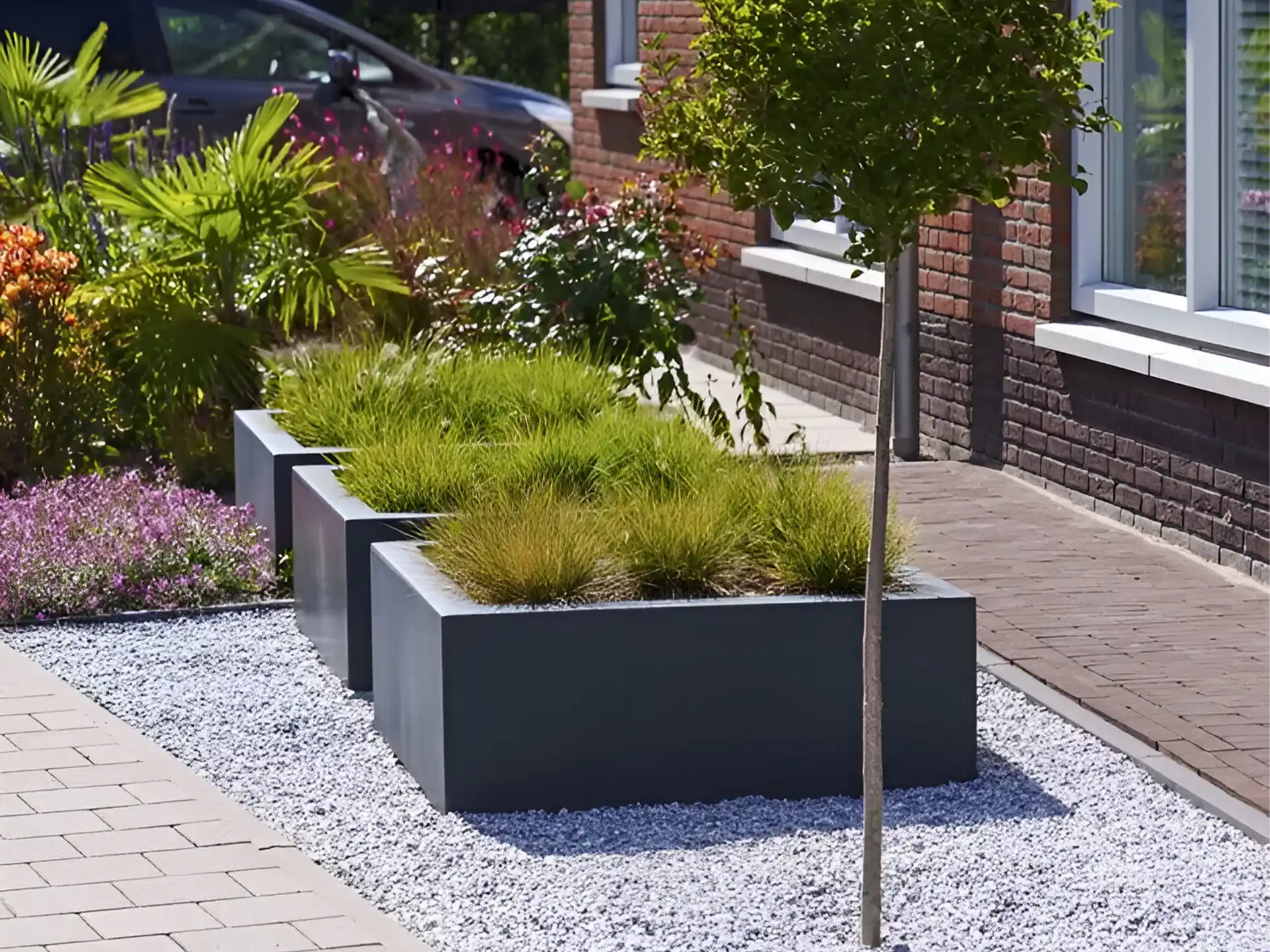 Adezz Carrez polyester plantenbak in antraciet, geplaatst naast een oprit met groene beplanting.