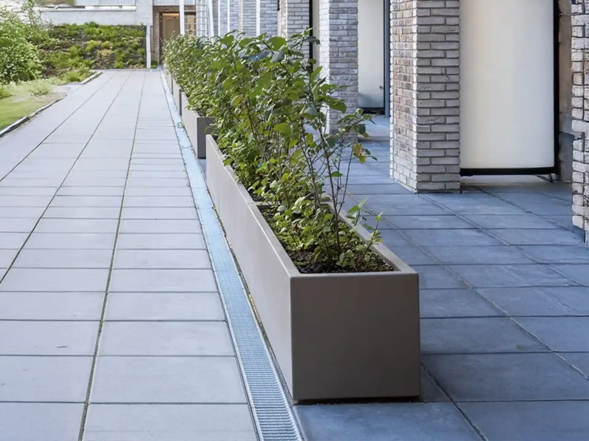 Taupe kleurige Adezz Carrez polyester plantenbak met wielen, geplaatst langs een moderne gevel met beplanting.