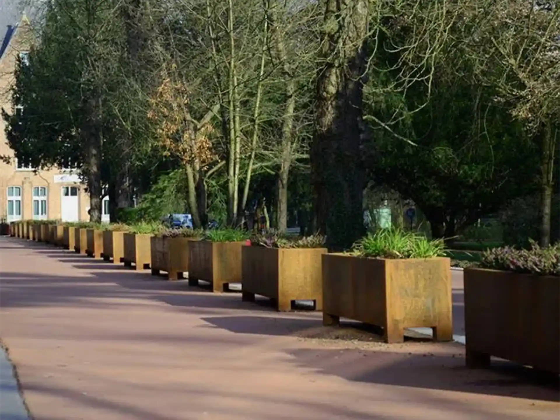 Meerdere Adezz Carrez Cortenstaal Plantenbakken Rechthoek met poten en bodem langs een straat in een groene omgeving.