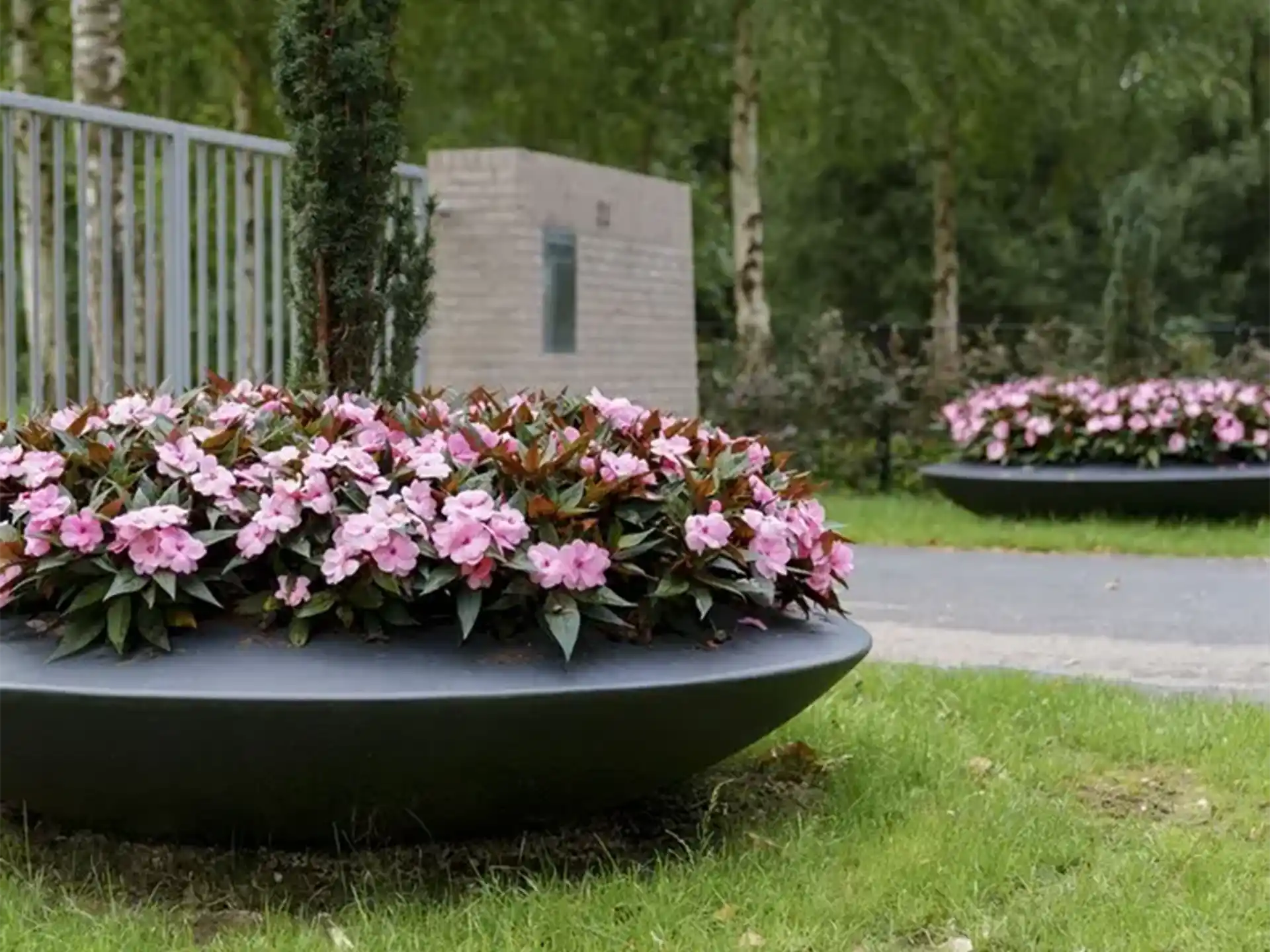 Adezz Ceder polyester plantenbak rond schaalmodel in zwart, gevuld met roze bloemen in een tuin.