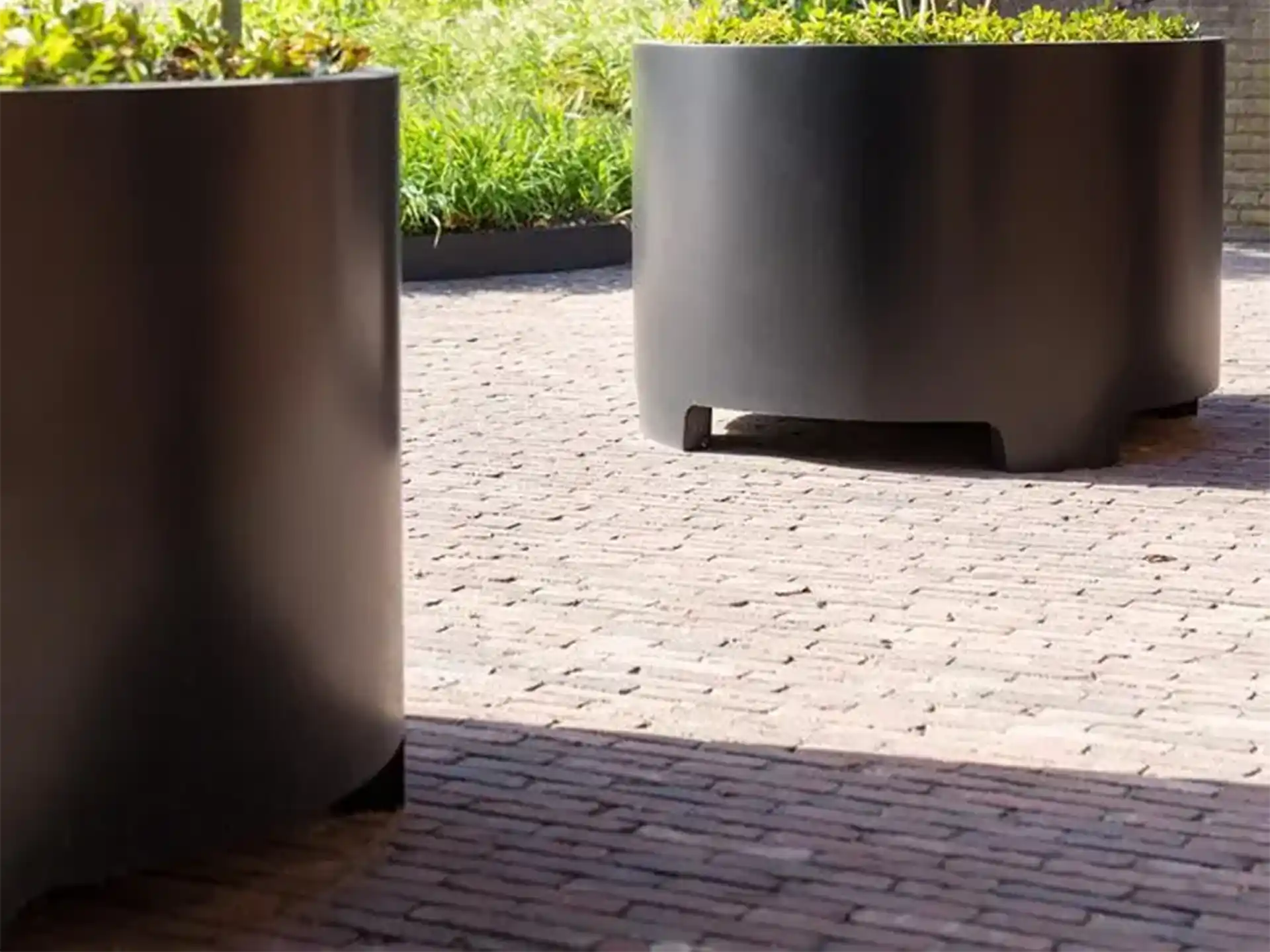 Zwarte Adezz Circum aluminium plantenbak met moderne poten op een terras met bestrating.
