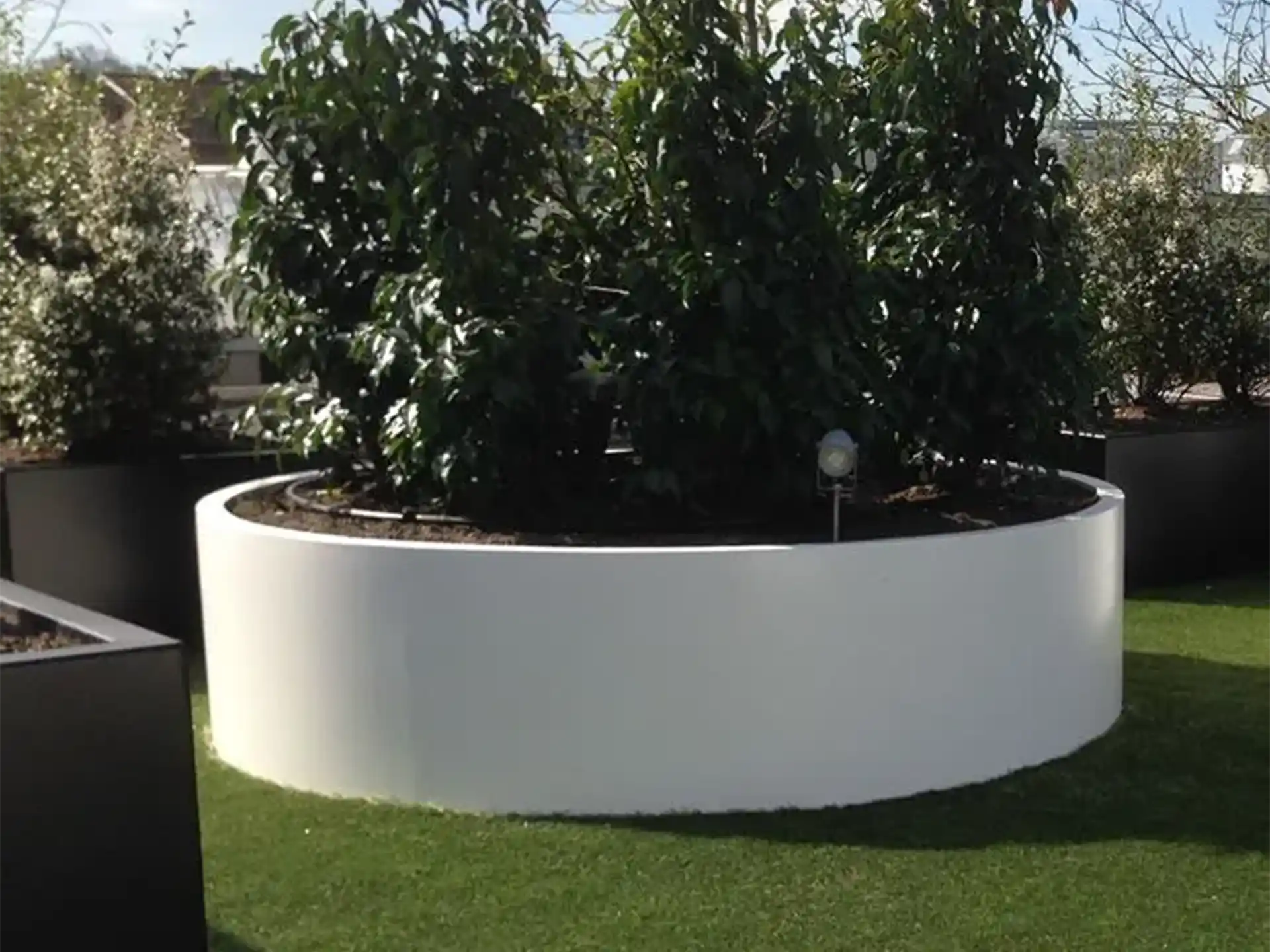 Grote ronde Adezz Circum aluminium plantenbak zonder bodem met meerdere struiken op een gazon.