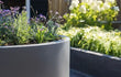 Adezz Circum polyester plantenbak rond recht in grijs geplaatst op een terras met bloemen en planten.