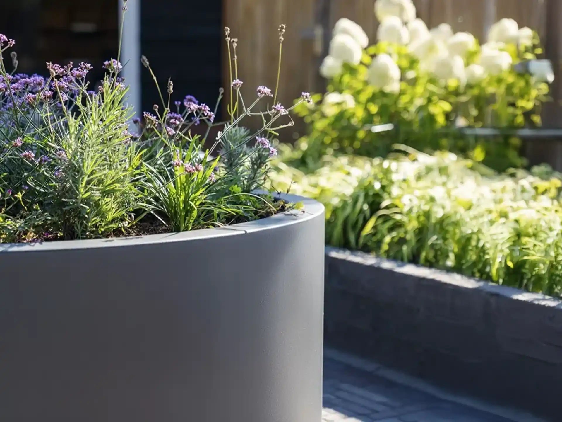 Adezz Circum polyester plantenbak rond recht in grijs geplaatst op een terras met bloemen en planten.