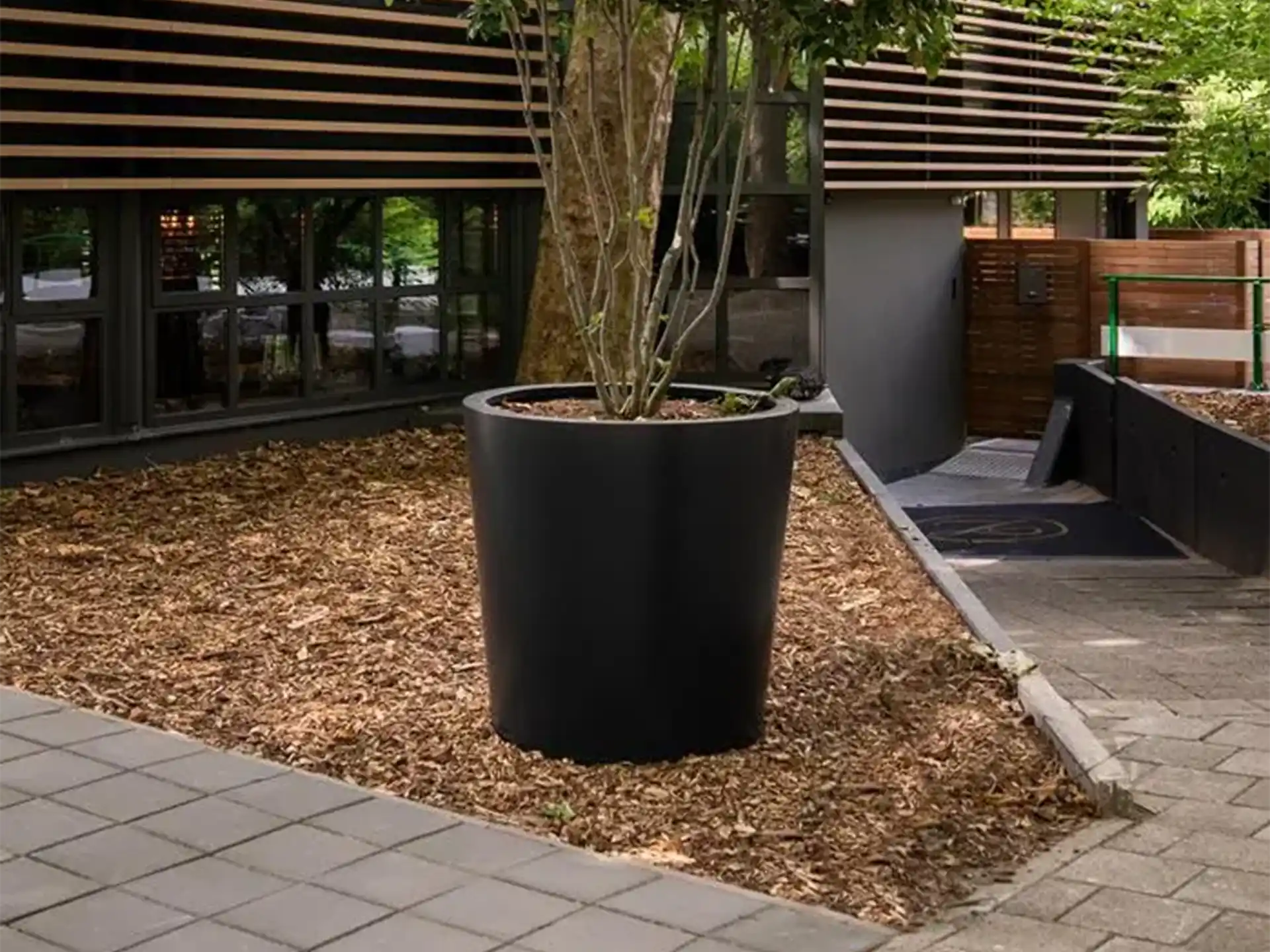 Grote Adezz Conic aluminium plantenbak rond met boom, geplaatst in een strakke tuin naast een modern gebouw.