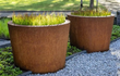Twee Adezz Conic Cortenstaal Plantenbakken Rond met bodem, beplant met siergrassen op een grindpad in de tuin.