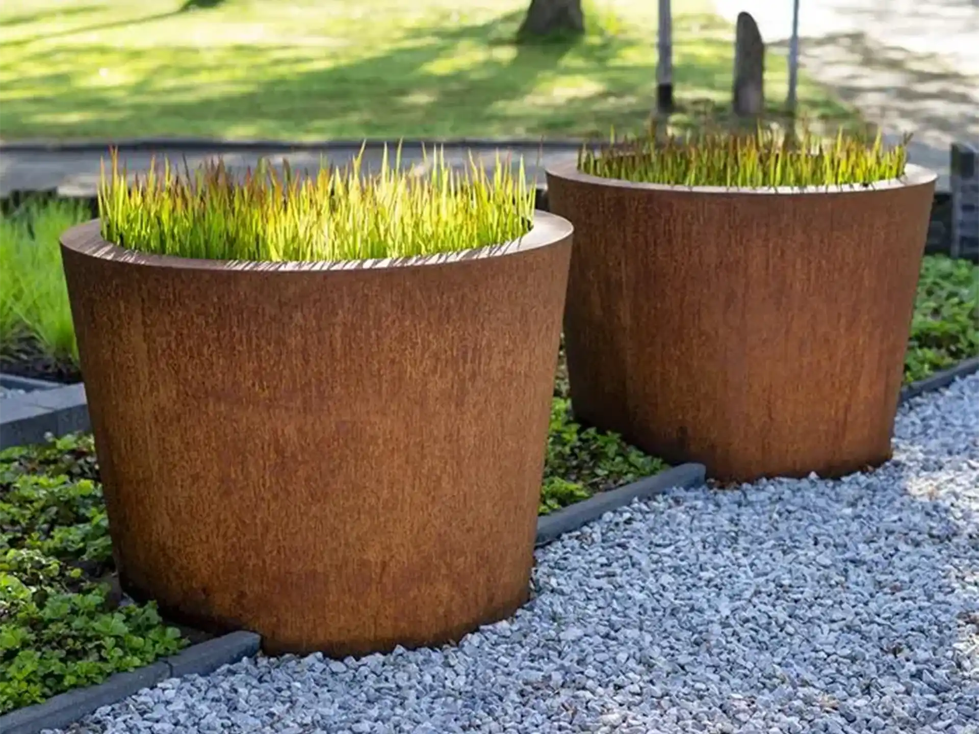 Twee Adezz Conic Cortenstaal Plantenbakken Rond met bodem, beplant met siergrassen op een grindpad in de tuin.