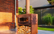 Adezz Forno Enok tuinhaard cortenstaal, geplaatst tegen een huisgevel, brandend vuur met houtopslag eronder, luxe buitenhaard.