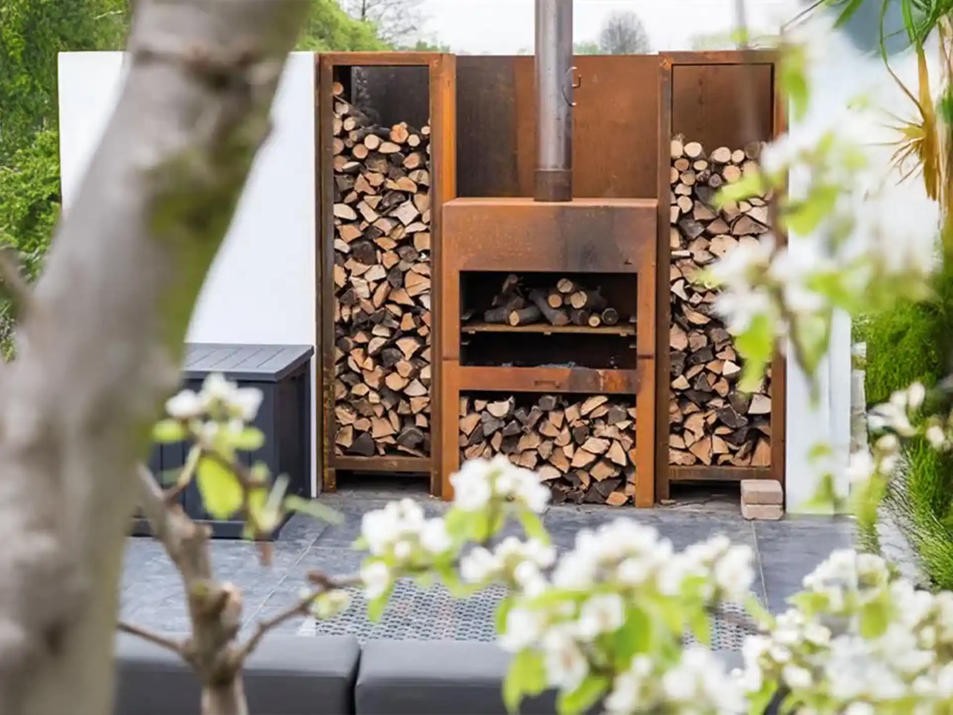 Adezz Forno Enok tuinhaard cortenstaal, stijlvolle tuinhaard met houtopslag, brandend vuur zichtbaar in moderne tuin.