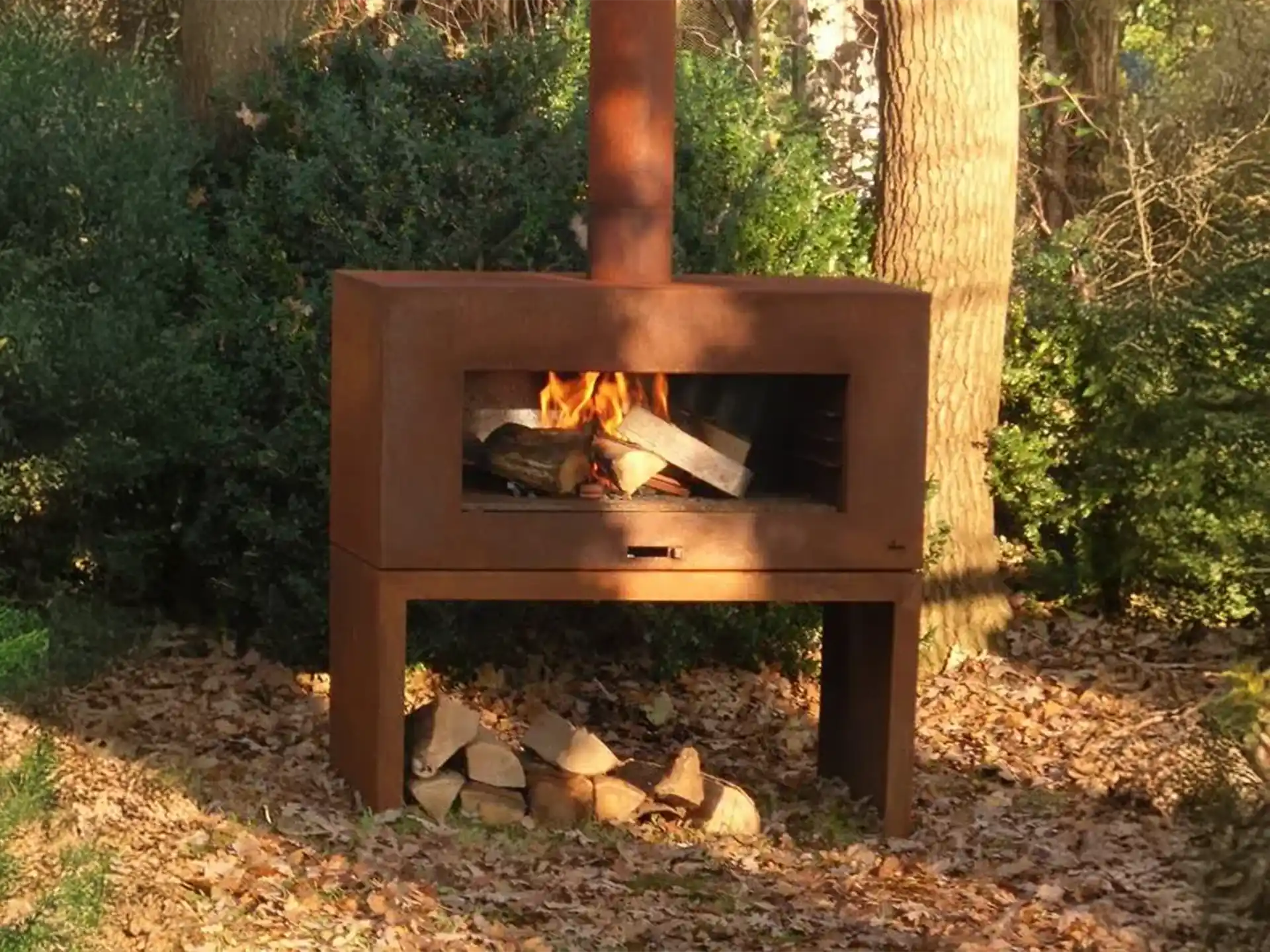Adezz Forno Enok tuinhaard cortenstaal, geplaatst in een groene tuinomgeving, brandend vuur zorgt voor gezellige sfeer.
