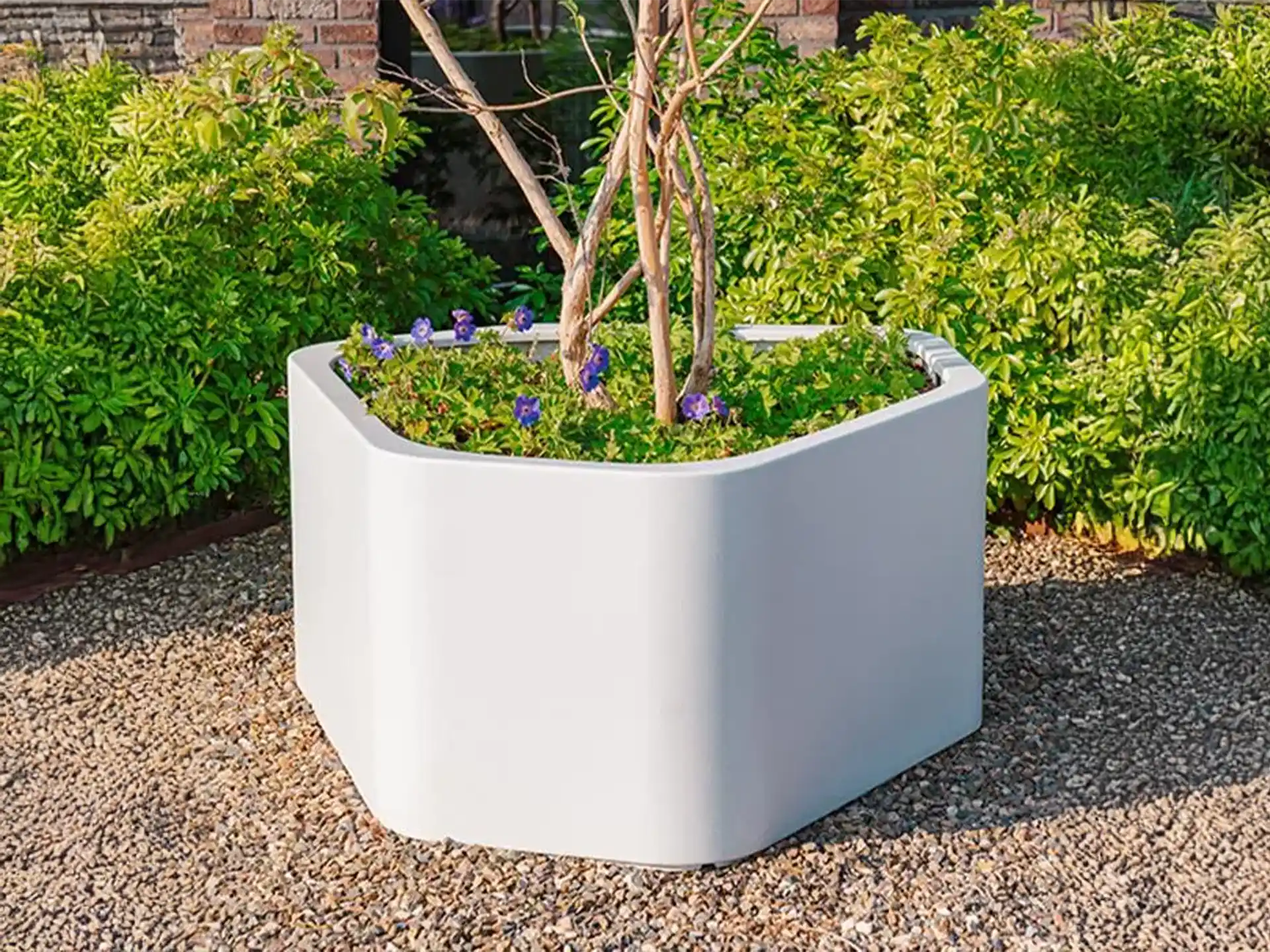 Adezz Shape polyester plantenbak rond hoeking model in wit, geplaatst in een tuin met grind en kleine boom.