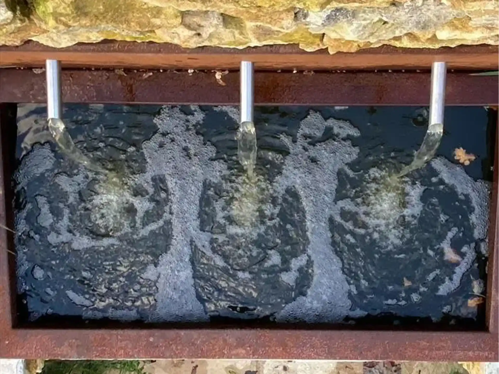 Close-up van drie waterstralen uit een cortenstaal Adezz vijvermuur, geplaatst op vijver met pomp, rustgevend waterornament in tuin.