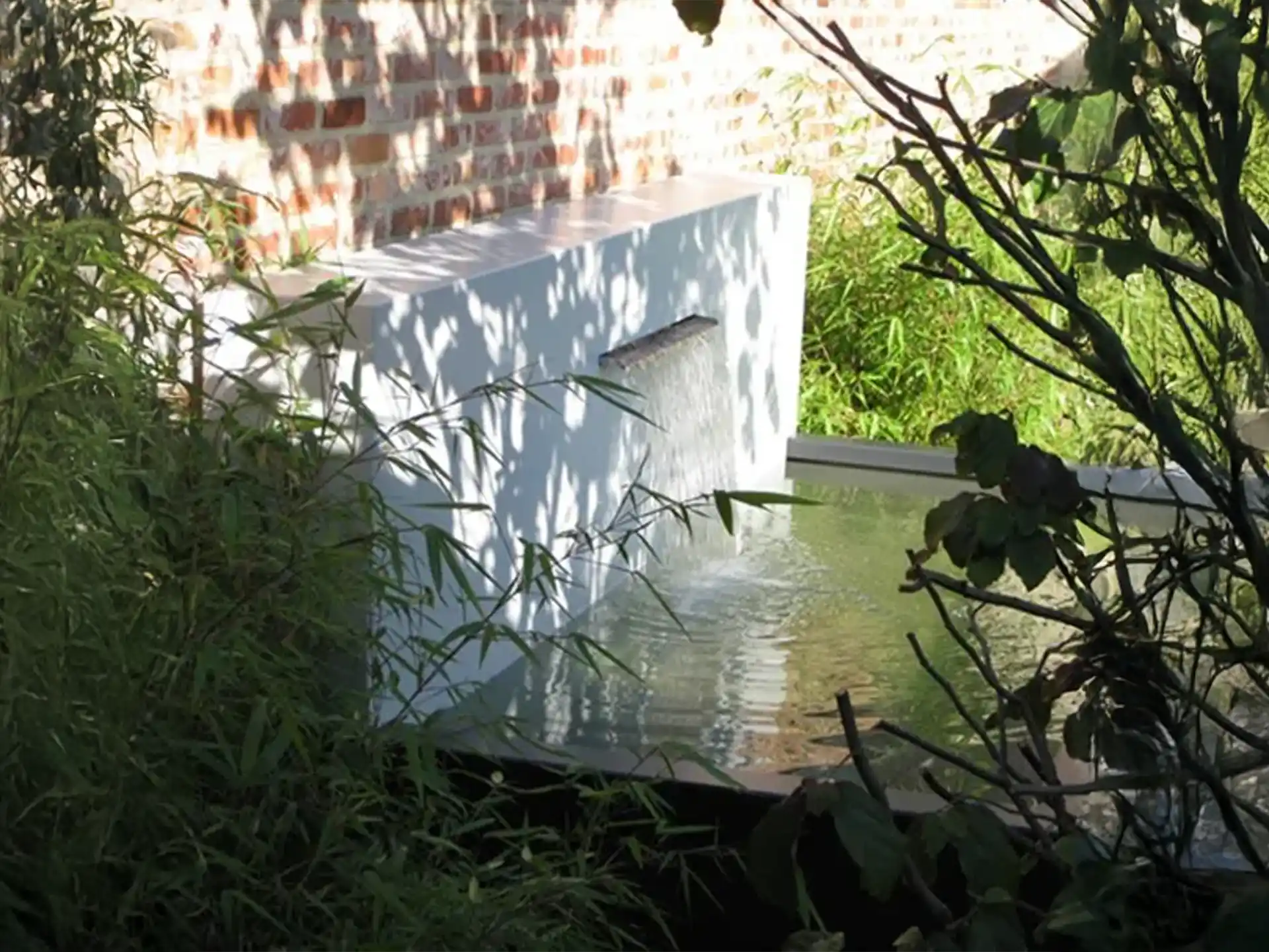 Adezz vijvermuur gemonteerd op aluminium vijver, waterornament met brede waterval, sfeervol in een groene tuin.