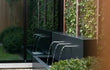 Sfeervolle tuinopstelling met een vrijstaande Adezz vijvermuur van aluminium, voorzien van 3 pijpjes en pomp, geplaatst langs een moderne gevel met glas en een houten schutting, waarbij waterstromen elegant in de vijver vallen.